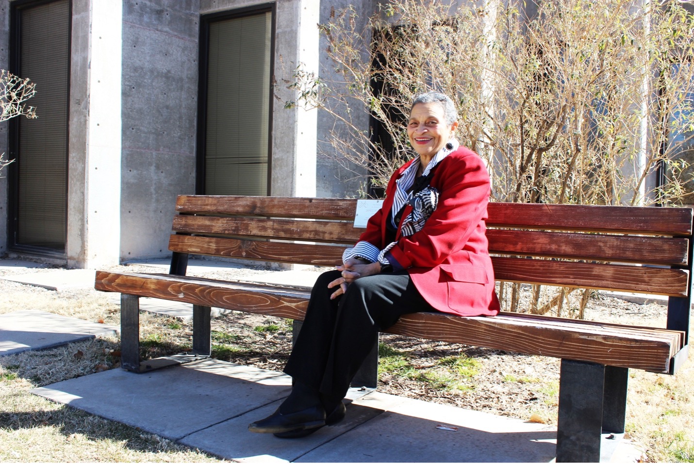 Marsha Hardeman, older woman of color sitting on a wooden bench in front of a concrete building