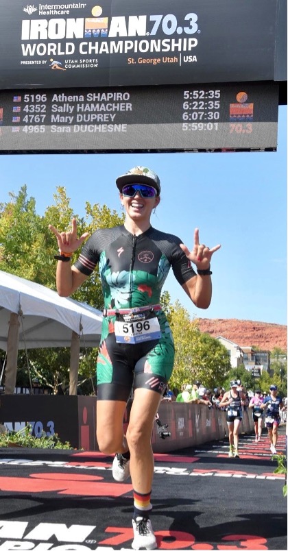 Athena Shapiro, young woman wearing athletic clothing and sunglasses smiles and she runs across the finish line