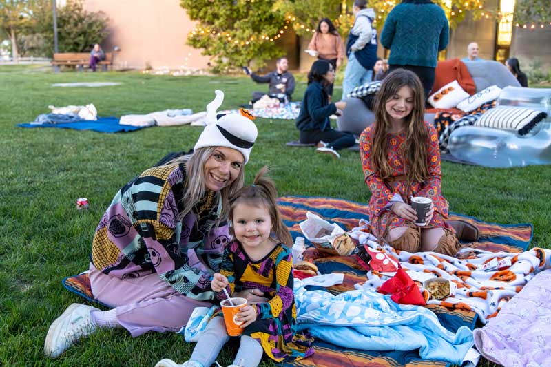 A mother and her daughters sitting on a blanket eating snacks